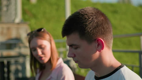 man in focus glances at woman beside him, blurred background highlights their interaction, with trees and iron railing enhancing the green hill backdrop