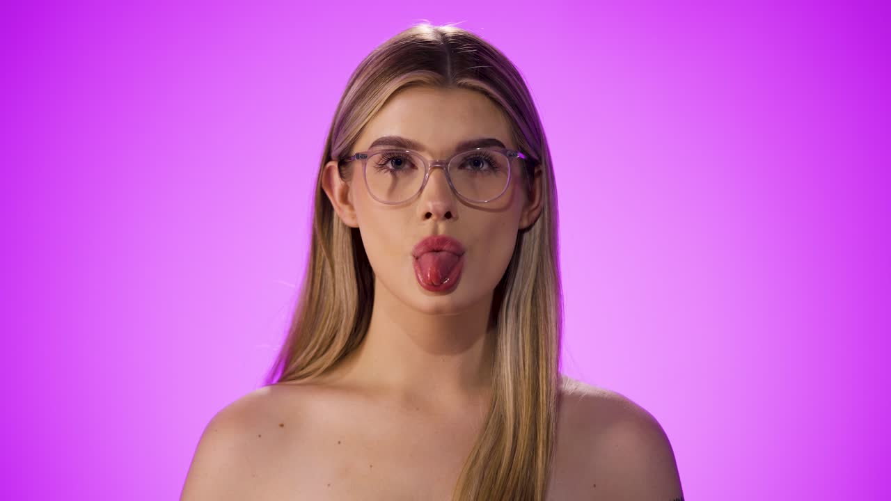 Medium close up shot of a young pretty woman wearing glasses and seductively sticking out her tongue into the camera in front of a purple background in slow motion