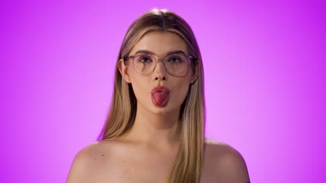 Medium-close-up-shot-of-a-young-pretty-woman-wearing-glasses-and-seductively-sticking-out-her-tongue-into-the-camera-in-front-of-a-purple-background-in-slow-motion