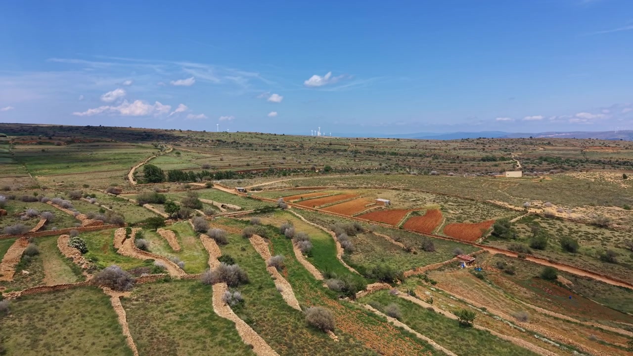 Terres agricoles en terrasses dans la province d'Aragon, Espagne, avec des champs de terre rouge, des broussailles sèches, des murs en pierre et des éoliennes à l'horizon sous un ciel bleu.