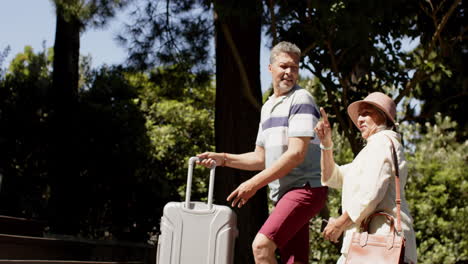 Happy-diverse-senior-couple-walking-with-luggage-to-a-house-in-sunny-outdoors