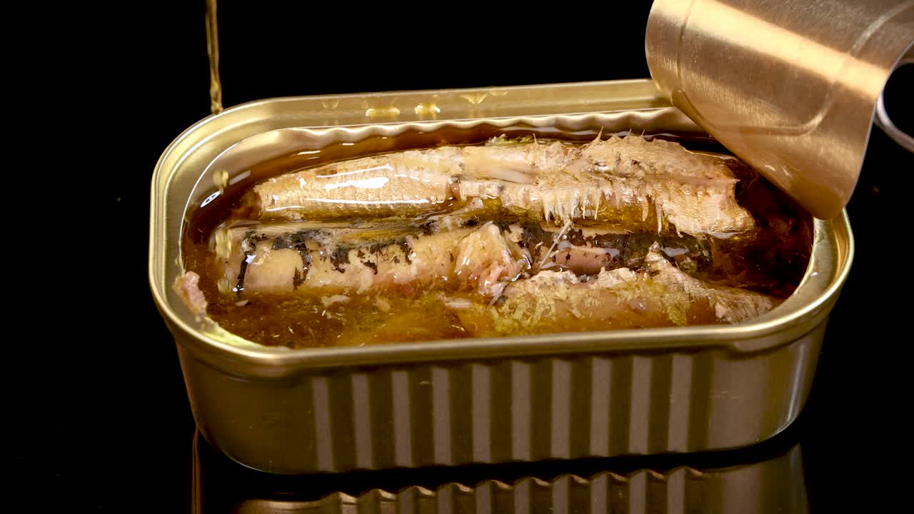 A metal fork lifts a sardine from an opened tin can filled with oil, under bright studio lighting with a black background and minimal reflections