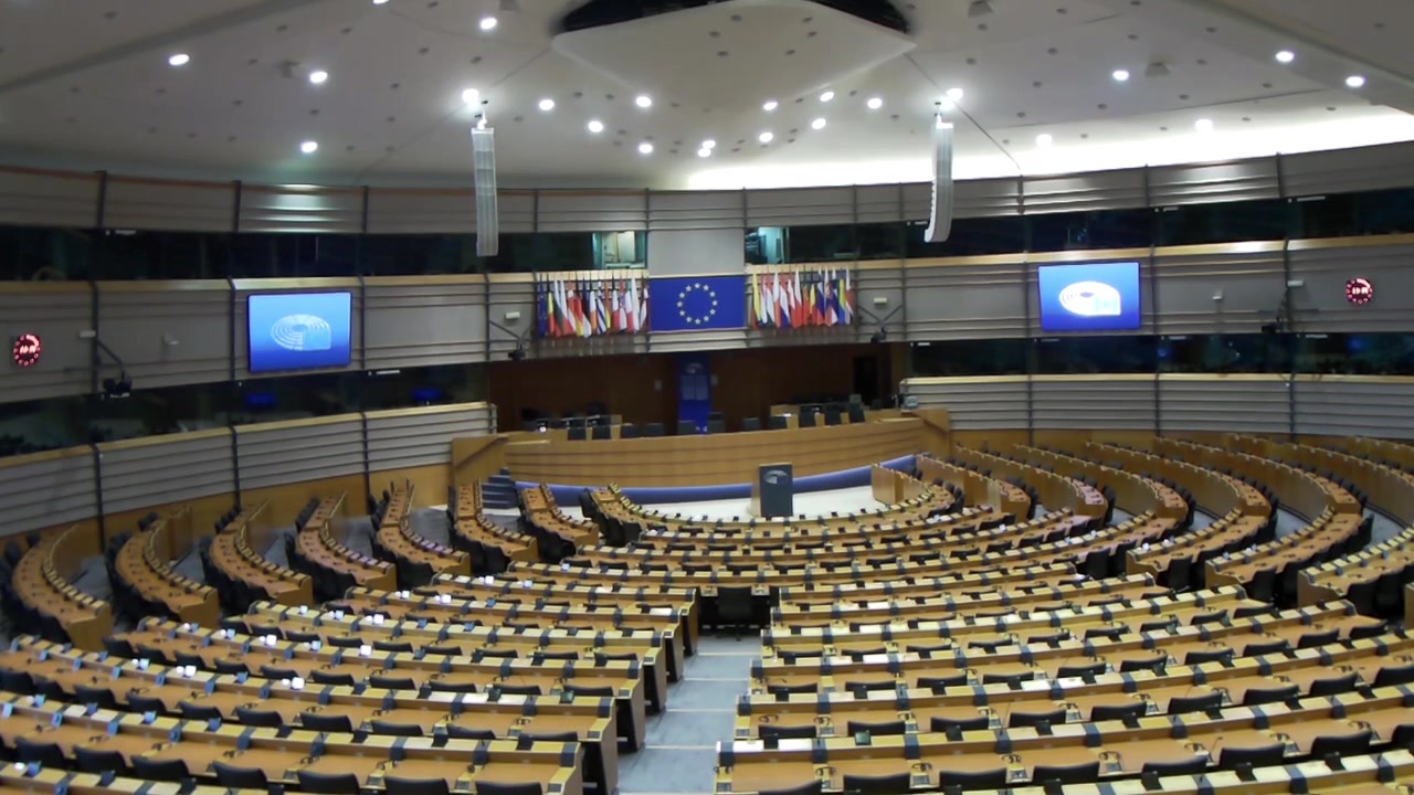 Aula del Parlamento Europeo a Bruxelles con posti vuoti e bandiere dell'UE, ripresa in grandangolo.