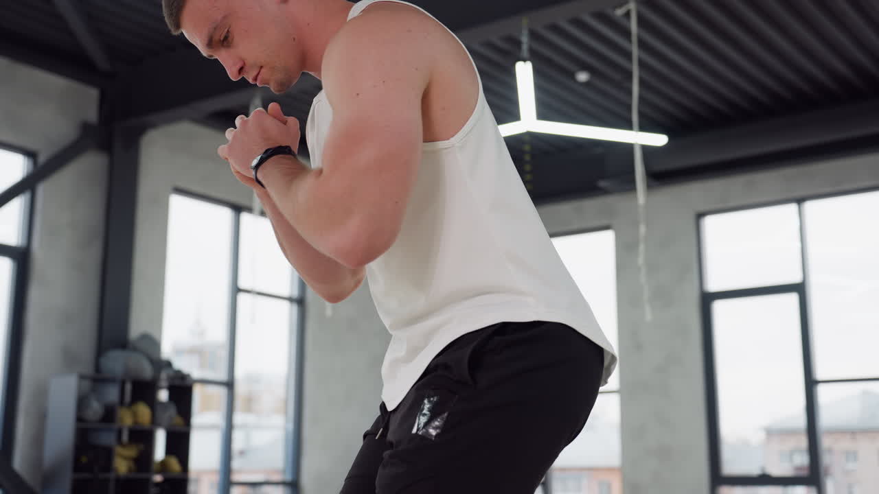 Gym enthusiast clapping hands while lunging forward for dynamic swatting exercise in bright studio, wearing white tank top and black shorts, large windows and modern suspension straps overhead