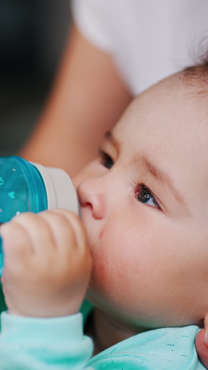 Lovely toddler with stylish hair drinking water from a bottle. Mom's hand is holding a bottle for her kid. Close up. Vertical video