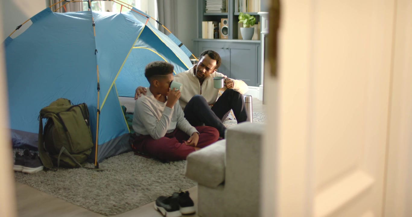 African American father speaking, touching shoulder comforting teen son sipping mug by tent at home