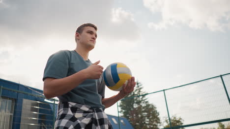 young man with plaid cloth around waist rotates volleyball in hand while standing outdoors, light creates a glowing effect around his shoulder, with trees and open space in background