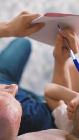 young bald father and little daughter student lie on floor of house, draw something pencil together in notebook, on distance learning, performs school homework on vacation