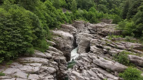 Drone-view-of-wild-water-flowing-under-rocks,-surrounded-by-forests-and-mountains-in-the-natural-beauty-of-Switzerland