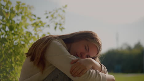 lady sits on grassy ground, arms folded over her knees, head resting on her hand, appearing deep in thought with sunlight softly illuminating her, the blurred background includes greenery