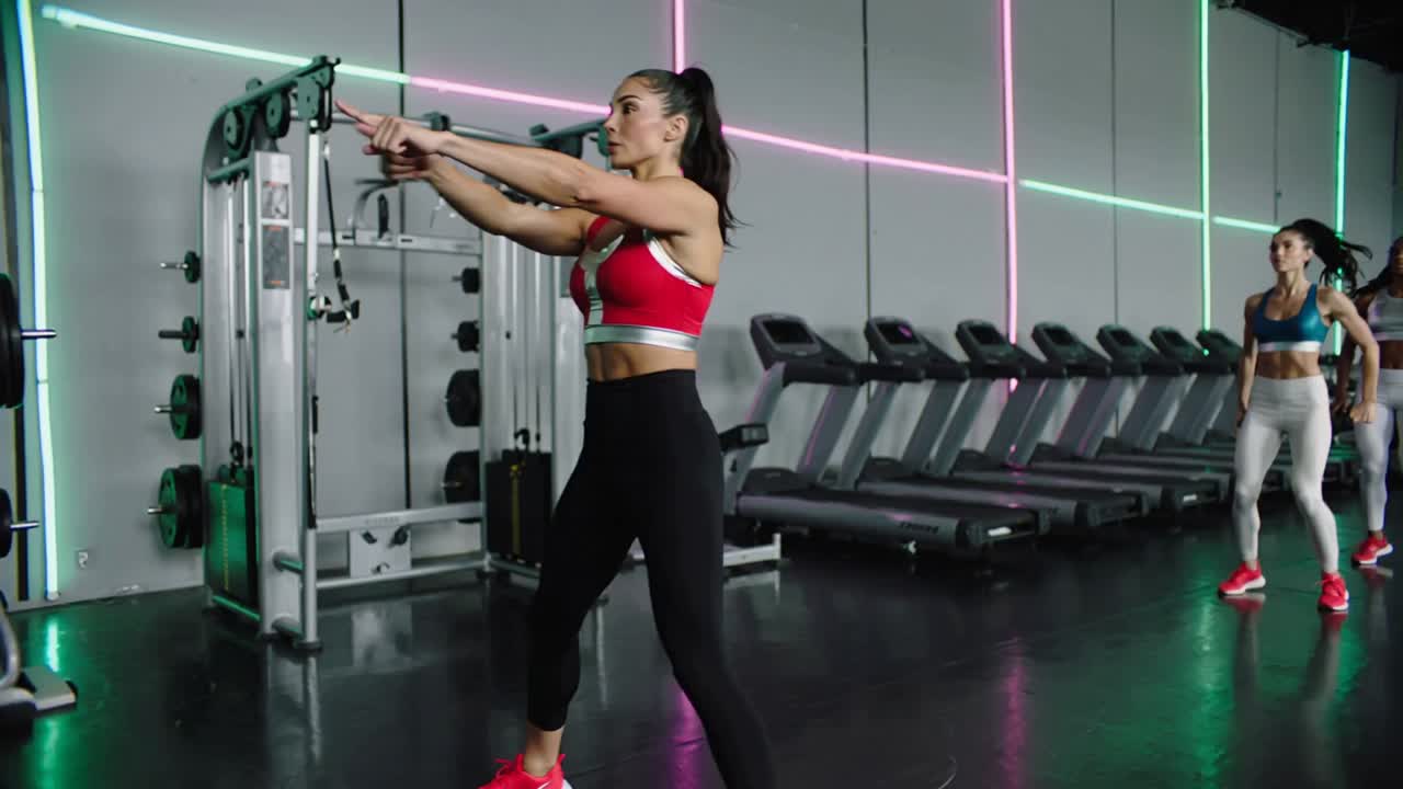 Demonstrating instructor wearing red sports bra cueing participants marching in gym with treadmills