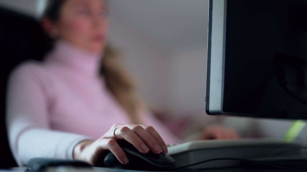 A woman focuses on her work in a comfortable home office. She uses a computer and mouse in a well-lit space, reflecting her dedication to completing tasks late at night
