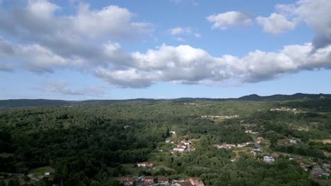 Drone-descends-on-scattered-homes-spread-throughout-dense-forest,-tilts-up-to-cloudy-blue-sky