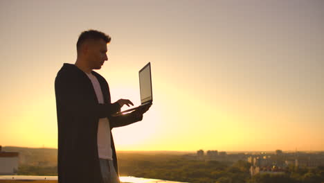 A-male-stockbroker-freelancer-stands-on-a-rooftop-at-sunset-with-a-laptop-and-types-on-a-keyboard-with-his-fingers-looking-at-the-cityscape-from-a-bird's-eye-view.