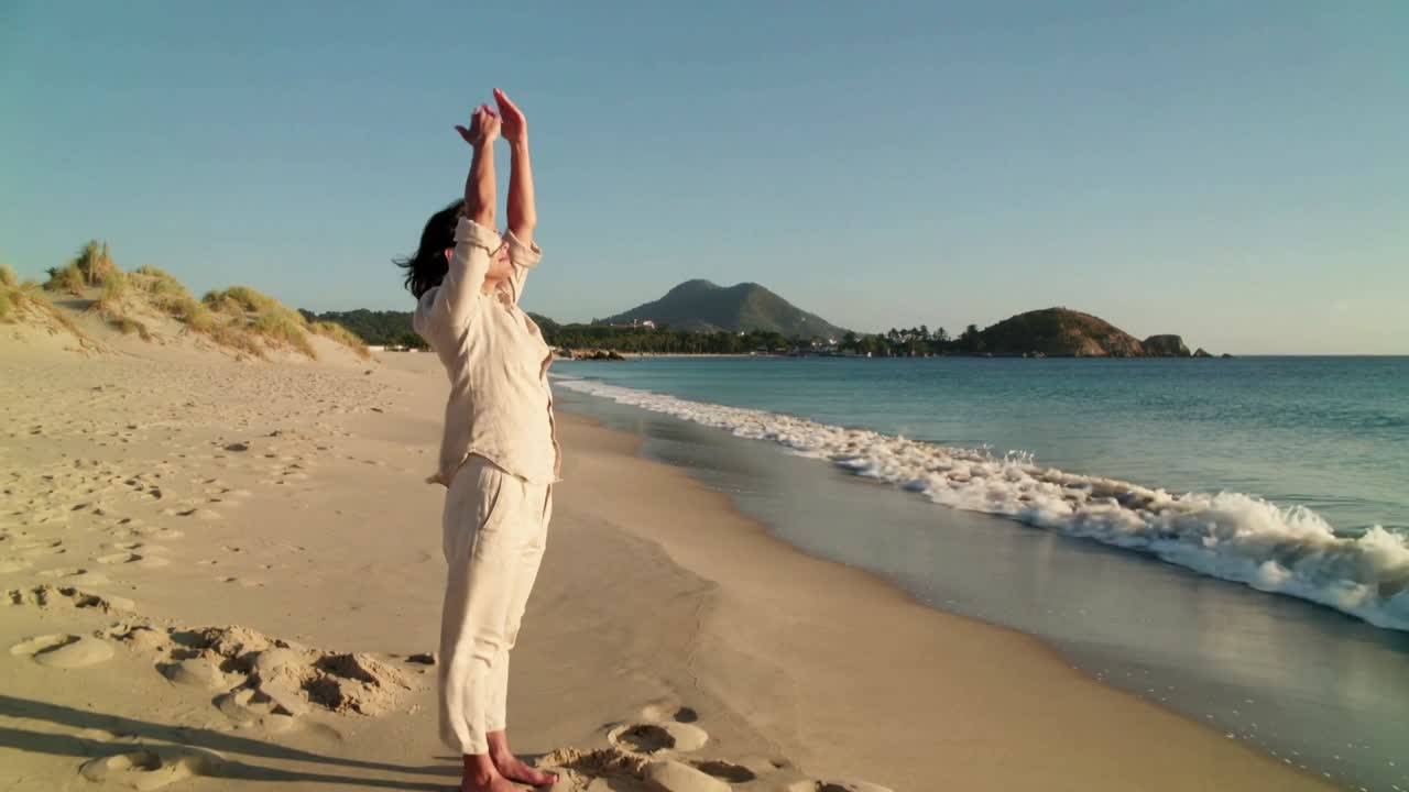 Uomo in lino che si allunga sulla spiaggia sabbiosa, sollevando le braccia sopra la testa mentre il sole gli illumina il viso; si vedono impronte.
