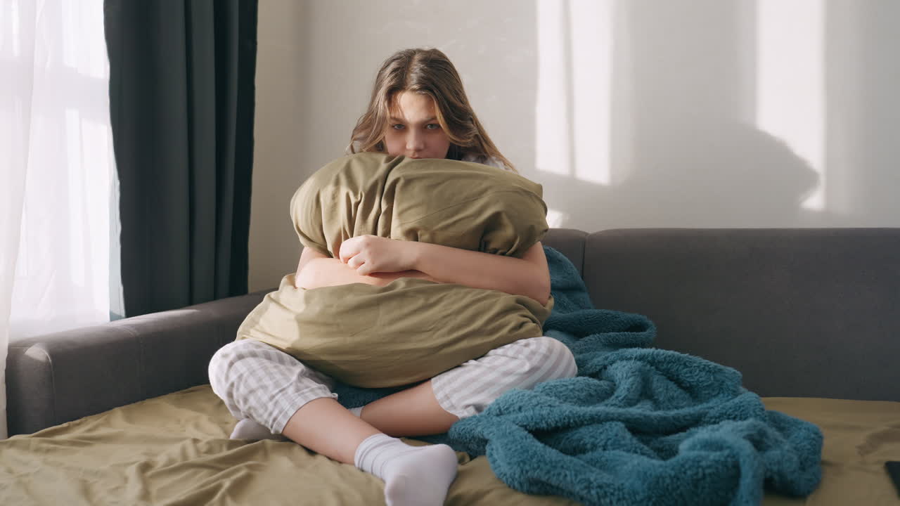 White Teenage Girl Hugging Pillow On Couch, Crosslegged With Cozy Blanket And Daylight Through Window, Anxious Expression And Self Soothing Posture, Casual Pajamas And Soft Texture Creating Intimate