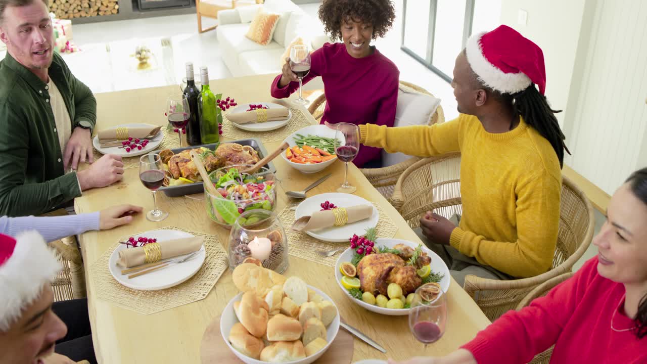 Diverse friends adjusting napkins, tapping hands, passing dishes, toasting wine at holiday table