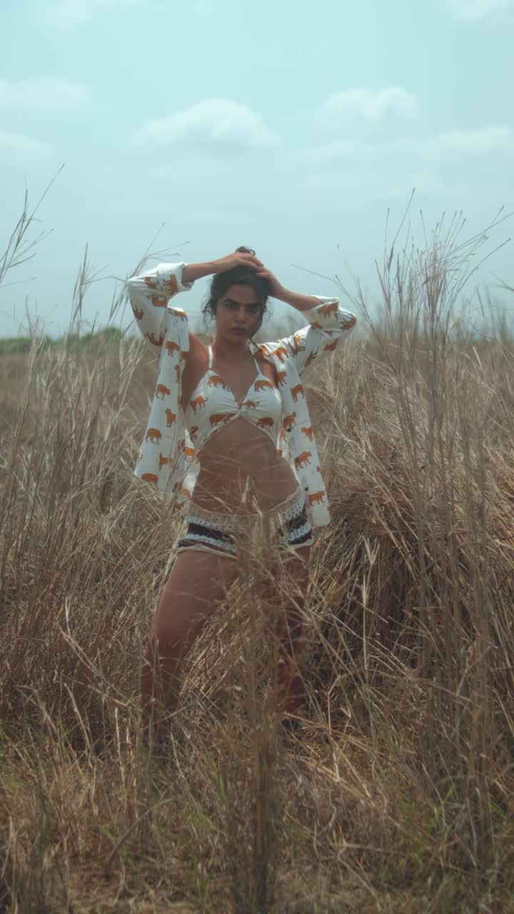 A young woman in a field of tall dry grass, posing in a stylish outfit under clear skies