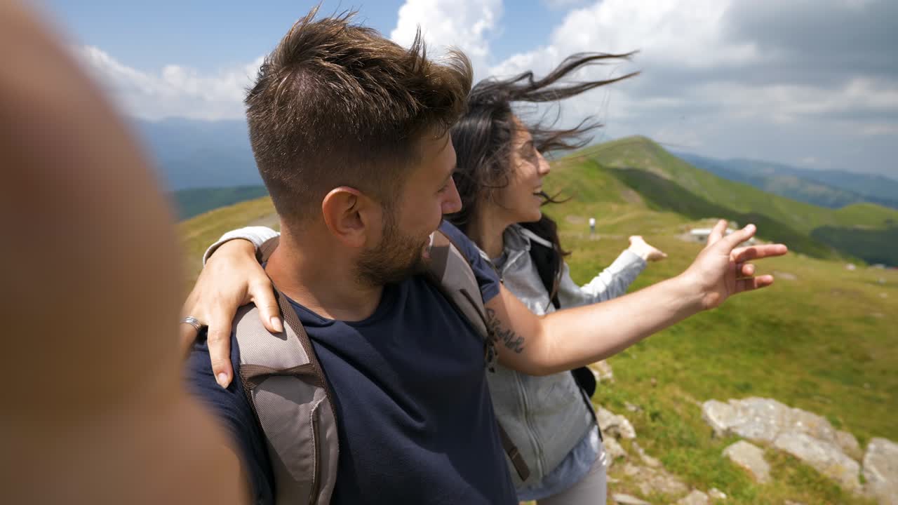 una pareja de turistas felices y despreocupados con mochilas está haciendo una llamada de video de selfie o tecnología a amigos o parientes que acaban de alcanzar un pico mientras caminan en medio de colinas rodeadas de naturaleza verde.