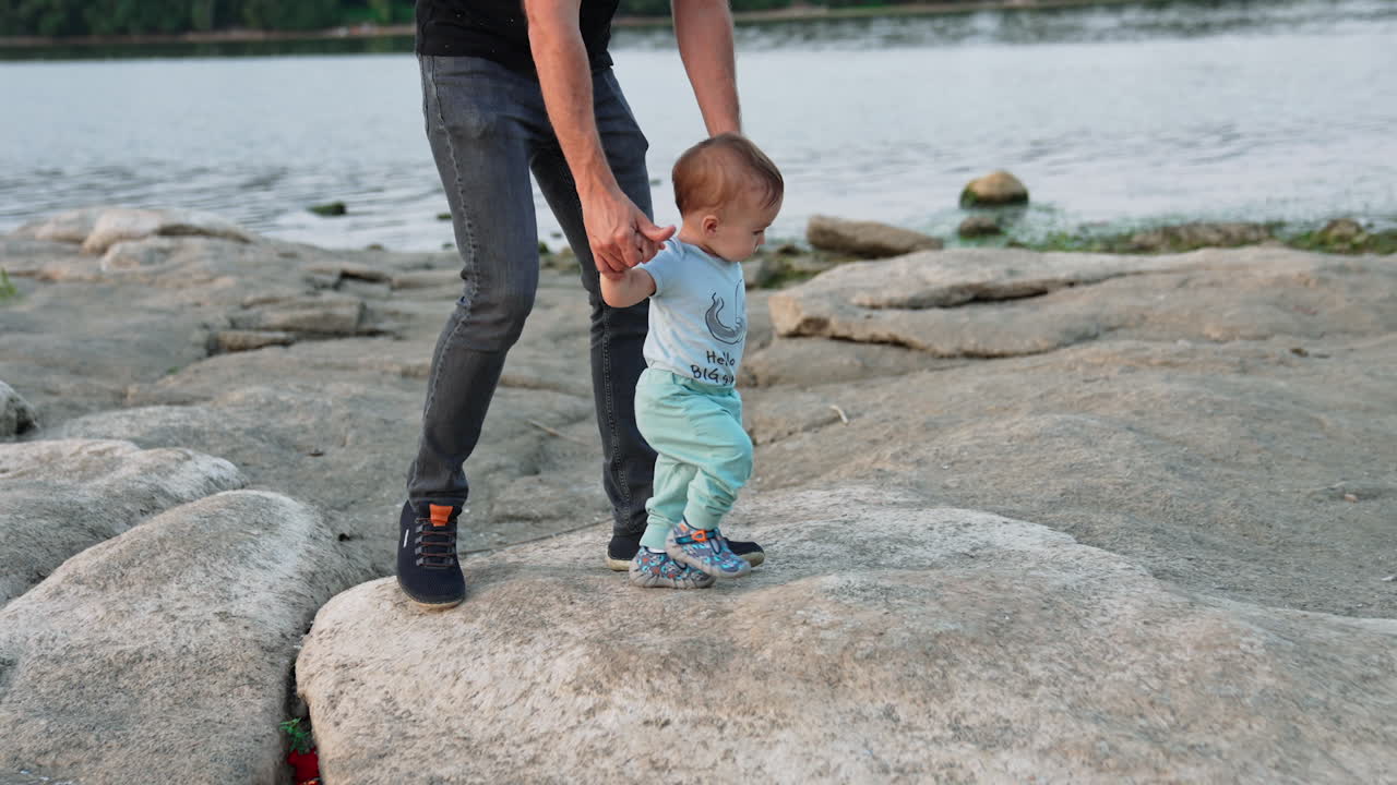 Little cute toddler stepping by the stones supported by his father. Loving dad teaching baby to walk in nature. River at backdrop.