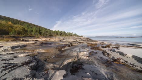 Die-Glatte,-Felsige-Küste-Verbindet-Sich-Mit-Einem-Sandstrand,-Der-Von-Seetang-Und-Algen-Bedeckt-Ist,-Während-Im-Hintergrund-Ein-Zeitraffervideo-Mit-Langsam-Rollenden-Wellen-Zu-Sehen-Ist