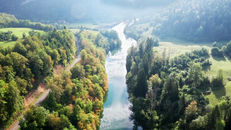 Route de campagne sinueuse longeant une rivière bleu vif, avec des forêts verdoyantes reflétant le soleil, vue aérienne par drone