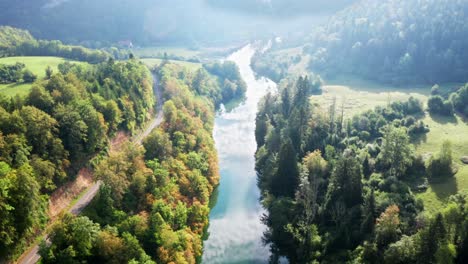 Kurvenreiche-Landstraße-Entlang-Eines-Strahlend-Blauen-Flusses-Mit-Sonnenreflektierten-Grünen-Wäldern,-Luftdrohne