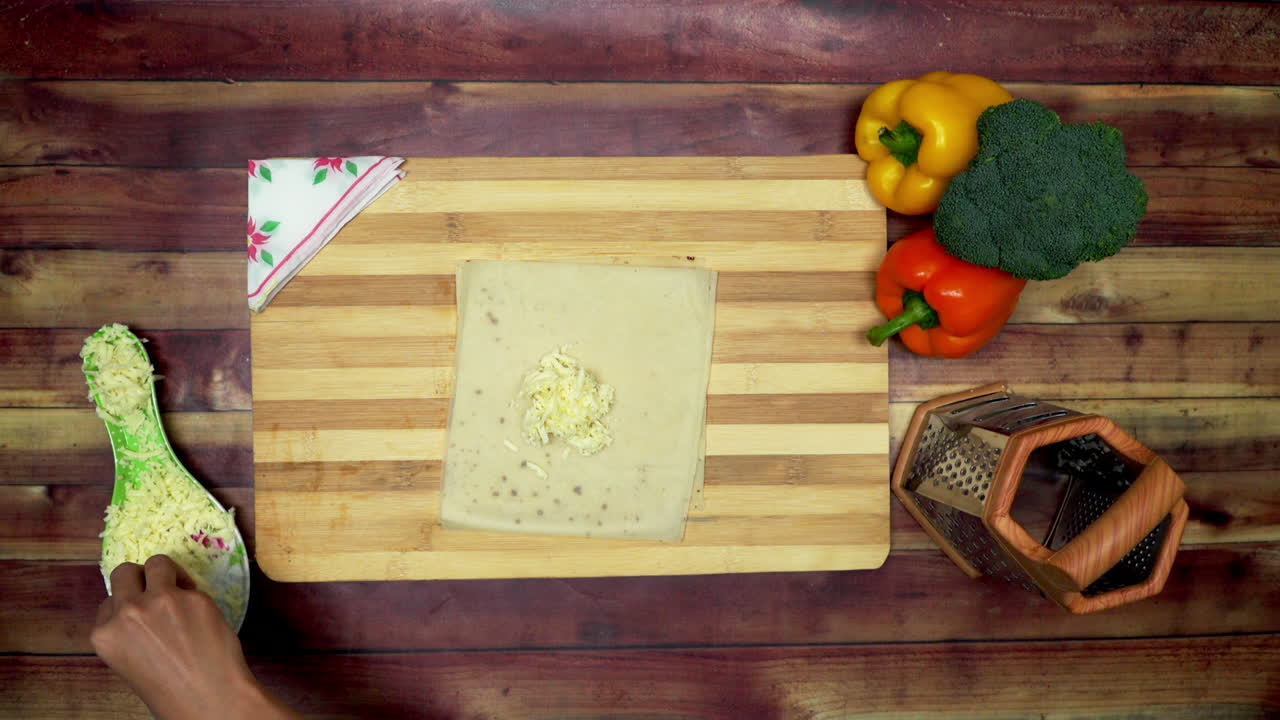 una vista superior de esparcir queso para el rollito de primavera, dos grandes pimientos amarillos y rojos, un rallador y un brócoli verde sobre la mesa