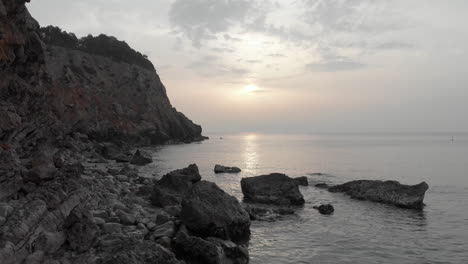 as-sun-rises-on-horizon-of-calm-sea,-low-tracking-drone-shot-glides-along-rocky-coastline