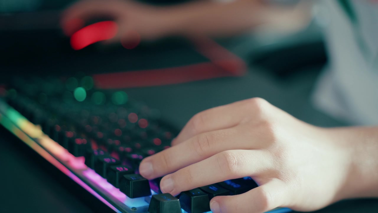 Close up of a teen hand pressing keys on a colorful backlit mechanical keyboard. Gamer playing computer game in a dark room, technology lifestyle and modern cyber entertainment, 4k.