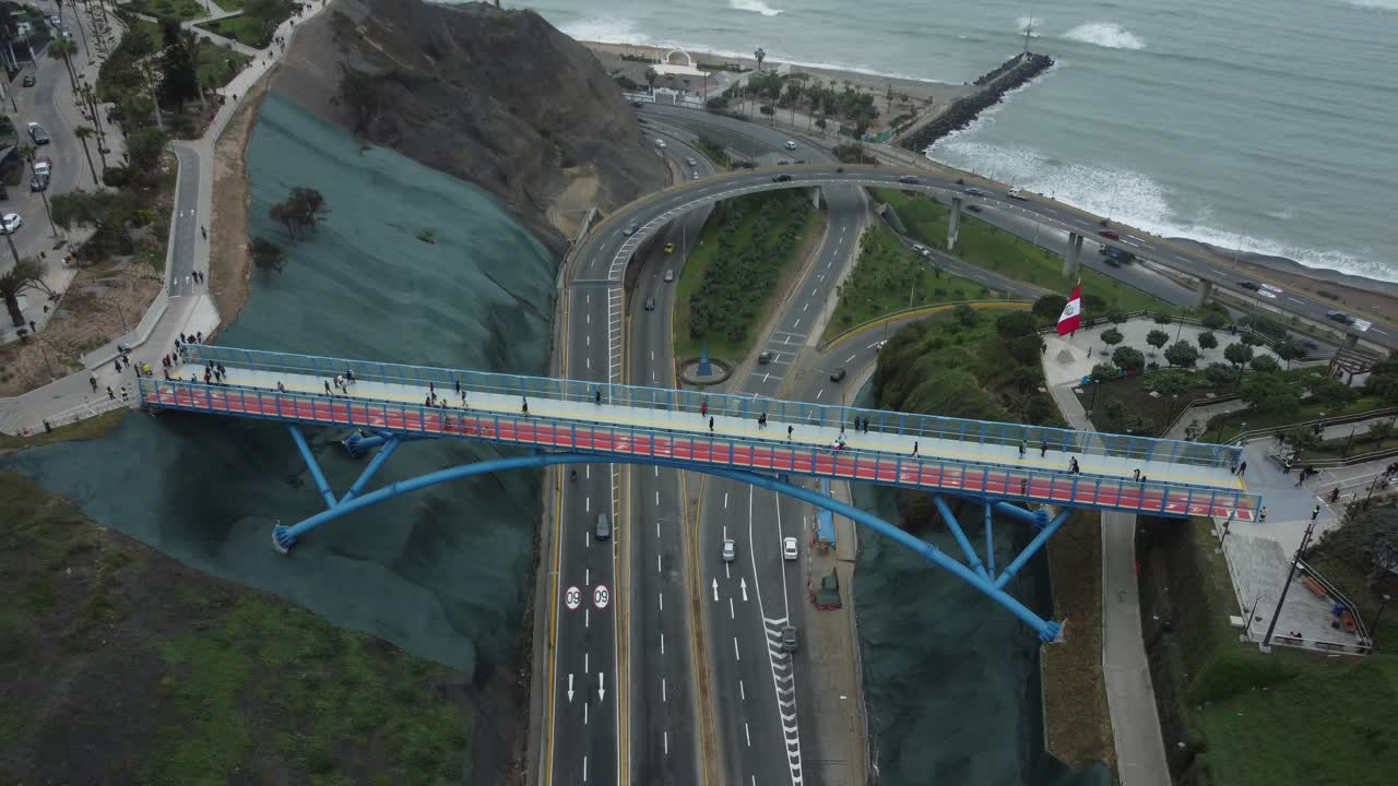 Aerial shot of a blue bridge above a street called "Bajada Armendariz" in Lima, Peru. The bridge connects two districts of Barranco and Miraflores. Drone flies backwards revealing the Peruvian coast