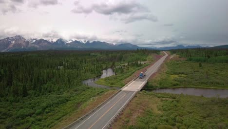 Großer-Sattelschlepper-Fährt-An-Einem-Bewölkten-Tag-über-Die-Jarvis-Creek-Brücke-Auf-Dem-Alaska-Highway-Mit-Spektakulären,-Rauen,-Schneebedeckten-Bergen-Im-Hintergrund,-Luftstatik