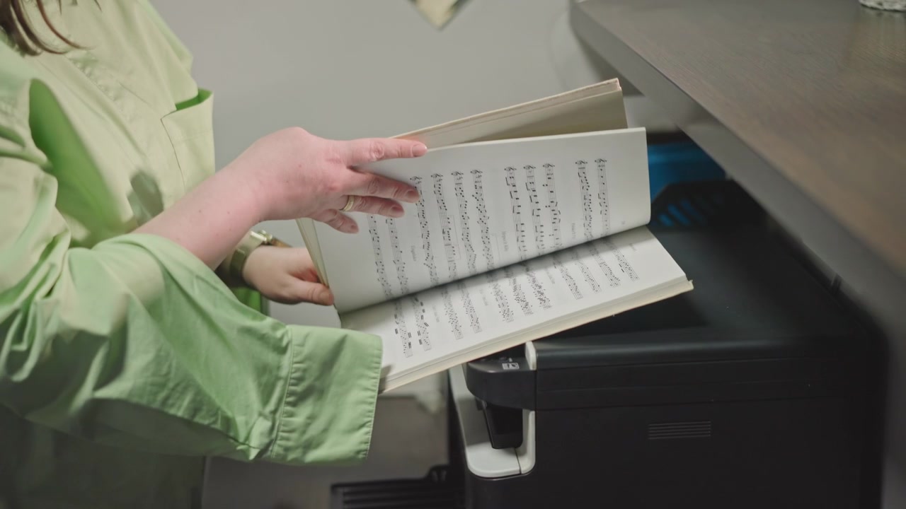 Music studio copy station preparing scores with hands feeding binder into printer and flipping pages to check notation, assistant in green uniform inspects document, compact machine and tray visible
