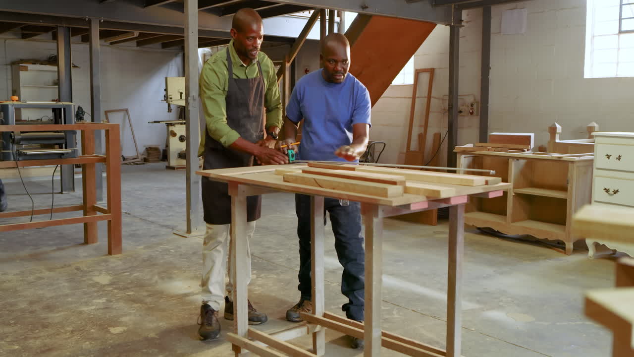 African American mentor pointing guiding apprentice measuring wood with tape marking cuts at shop
