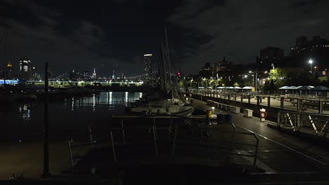 wide shot, filmed at night, of brooklyn docks, with several boats in the water and new york city skyline in background