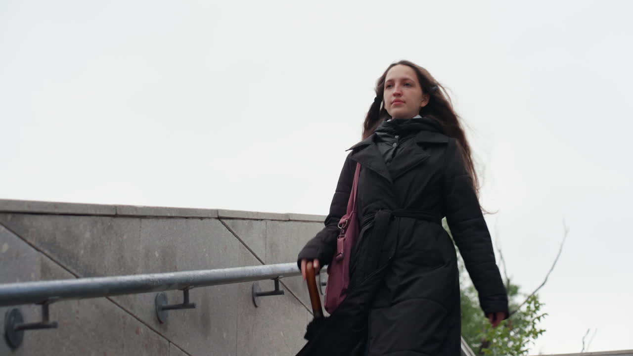 Donna bianca che cammina verso la fotocamera, cielo nuvoloso, cappotto nero, capelli lunghi al vento, borsa rosa di lato, passo costante lungo gradini di cemento, sguardo ravvicinato, espressione calma, pausa da pendolare, ringhiera urbana