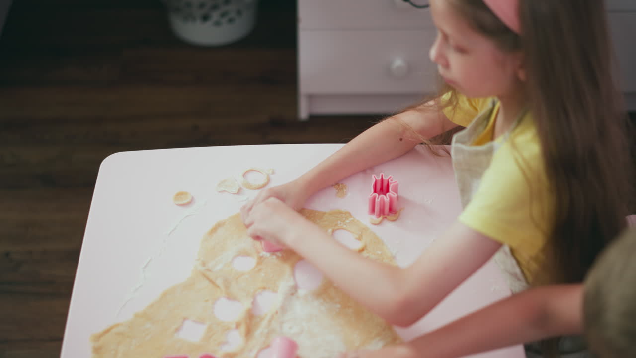 Overhead view of girl placing flour into shape cutter during baking activity while another kid focuses on dough, surrounded by cookie cutters and flour on white table