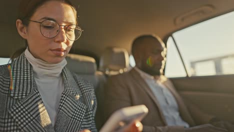 a confident girl businesswoman in round glasses in a gray jacket chats on her smartphone during her trip and business trip together with a male businessman with black skin in a brown jacket in a modern salon in a car on the road