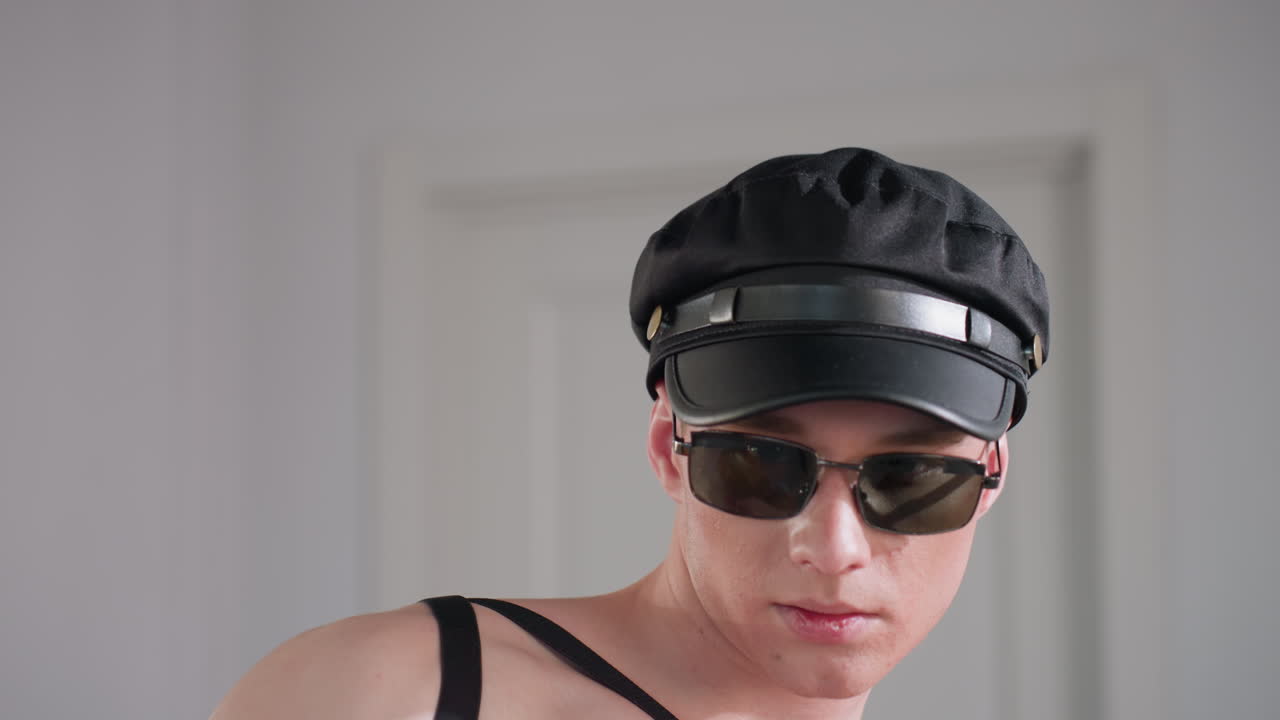 young man wearing black cap and sunglasses adjusts cap while licking thumb and posing stylishly inside bright white room with harness straps and closed door in background under soft lighting