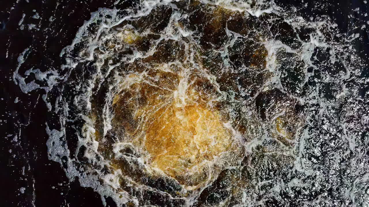 Turbulent, dark, bubbly water surface with an orange-brown central foam patch, photographed directly overhead, suggesting wake or boil, abstract, close-up