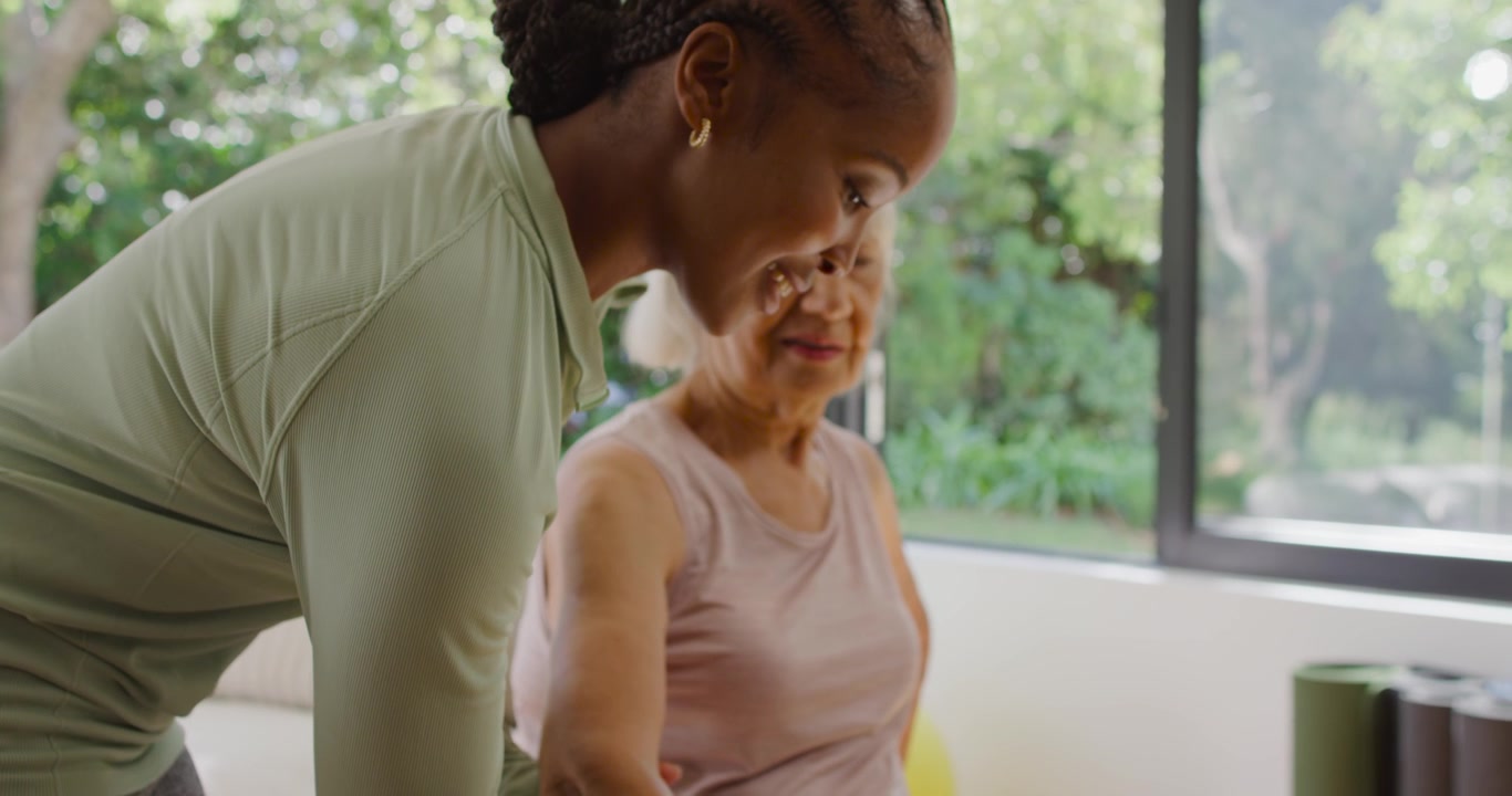 Mulher idosa levantando um halter roxo no sofá, enquanto um treinador afro-americano a guia manualmente para reabilitação.
