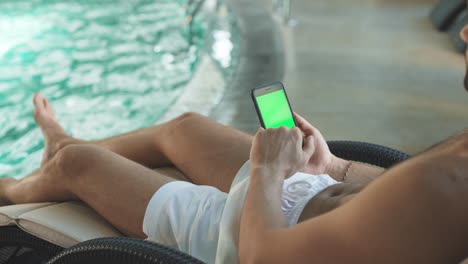 Manos-De-Hombre-De-Primer-Plano-Sosteniendo-Un-Teléfono-Móvil-Con-Pantalla-Verde-Junto-A-La-Piscina-Del-Hotel.