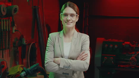 Portrait-of-young-Caucasian-woman-in-goggles-and-suit-smiling-at-camera-with-arms-crossed-in-workshop