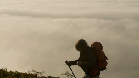 Senior-woman-with-backpack-climbing-mountain-with-trekking-sticks,-then-having-a-break-and-enjoying-the-view