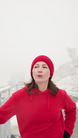coach exercising outdoors during winter holding iron railing for balance stretching leg back and forth surrounded by snowy ground foggy atmosphere distant river bridge background breath visible air