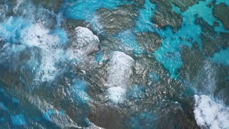 a drone captures a shot of a blue lagoon, showcasing the clear waters that reveal the underwater landscape of rocks, the waves crashing on the reef adding to the dynamic of the scene