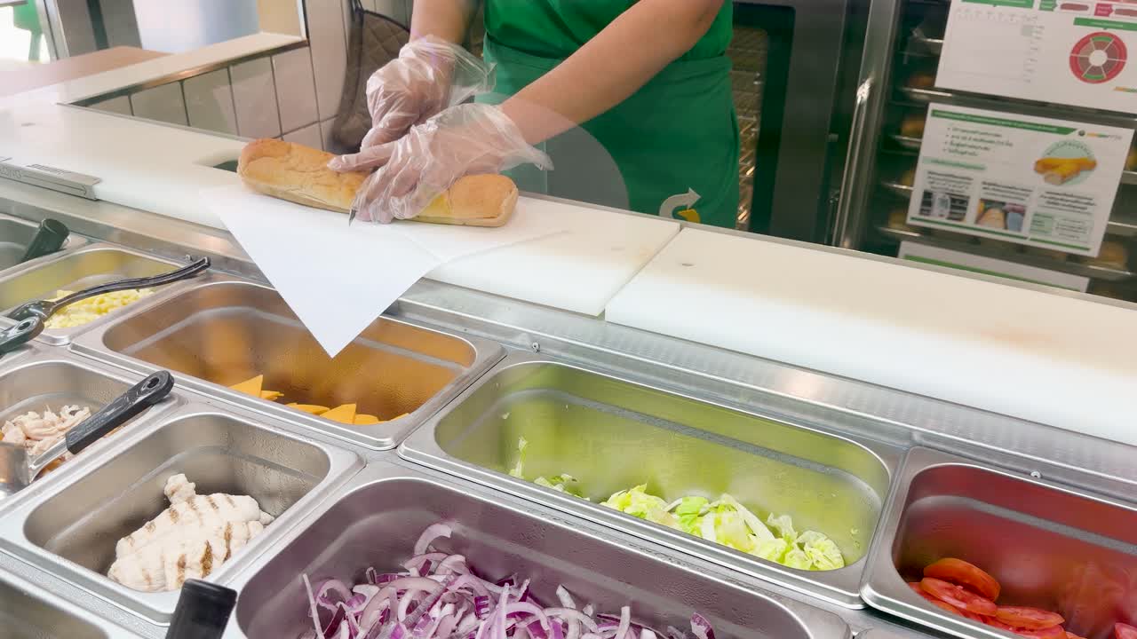 Un lavoratore che indossa guanti protettivi affetta un lungo panino da farcire a un bancone dei panini.