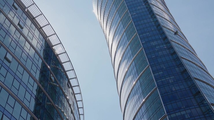 Alpinista industrial limpiando las ventanas de un rascacielos moderno con fachada de cristal curvado. Vista en ángulo contrapicado de un trabajador profesional realizando el mantenimiento de un edificio corporativo