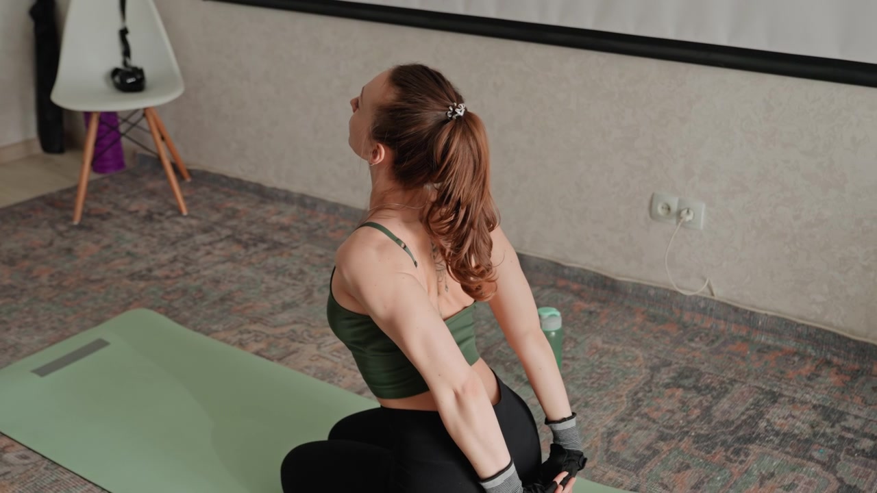 Caucasian woman house cooldown twist stretch, seated on mat performing shoulder and spine release with hands behind back, tattooed back in view, water bottle nearby for postworkout recovery