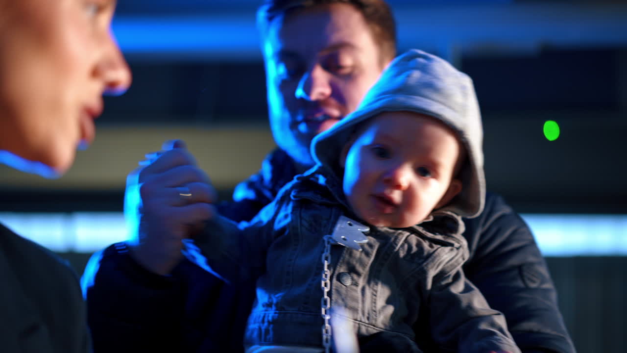 Thin silver candle on the cake is blown by a woman. Little kid in dad's arms tries to get the cake. Blurred backdrop.